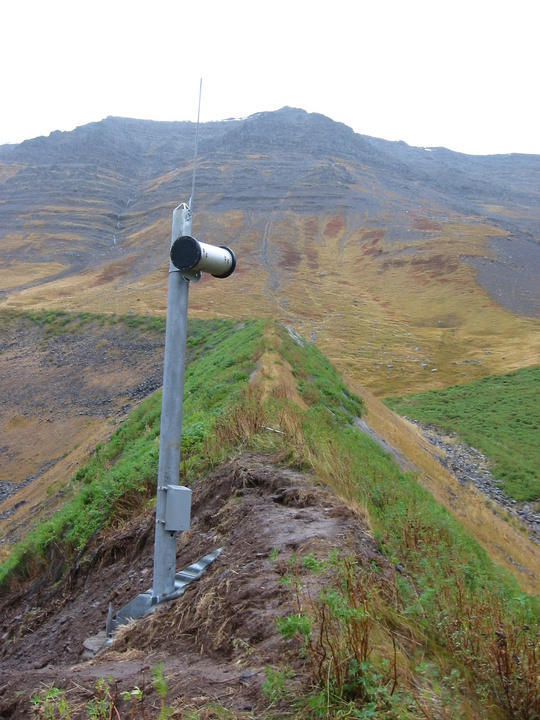 hólkur á stöng efst á grónum varnargarði, fjallshlíð í baksýn