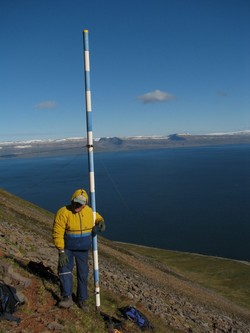 staðið við stiku í fjallshlíð, haf í baksýn