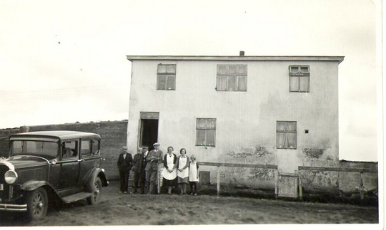 svarthvít mynd, lítill hópur fólks fyrir utan steinhús, gömul bifreið